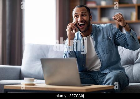 Cheerful Black man having phone conversation sitting at table with laptop in living room at home Stock Photo