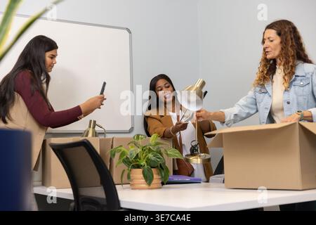 Diverse female coworkers unpacking boxes at meeting table, passing gold lamp, using smartphone Stock Photo