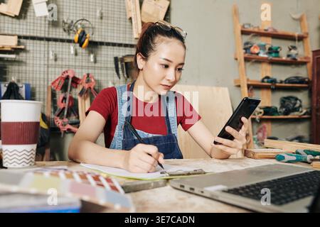 Asian female carpenter writing on planner reading text messages using smLifestylephone sitting at desk in woodshop Stock Photo