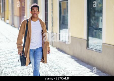 Smiling Black male carrying bag walking on street on sunny day Stock Photo