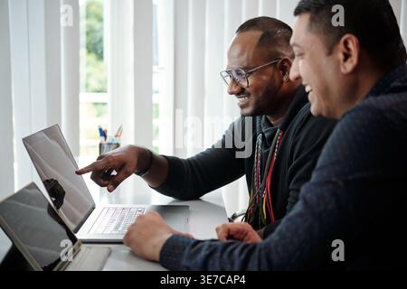 Asian male software developer checking programing code on laptop screen with male colleague while sitting at table in office Stock Photo