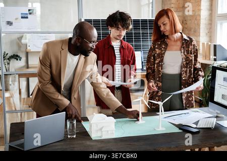 Diverse group of architects developing new project with models while standing at table with laptop and blueprints in office Stock Photo
