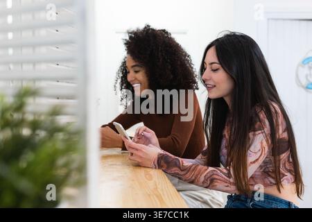 Mixed race and Hispanic female friends using their smartphones while sitting at wooden table in cafe Stock Photo