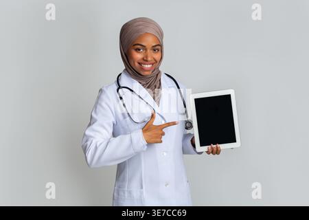 Black Muslim female doctor with stethoscope wearing hijab pointing finger on picture frame while standing against grey background in studio Stock Photo