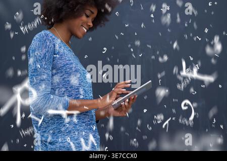 Smiling Black woman with afro using tablet standing against gray background with flying letters and symbols in studio Stock Photo