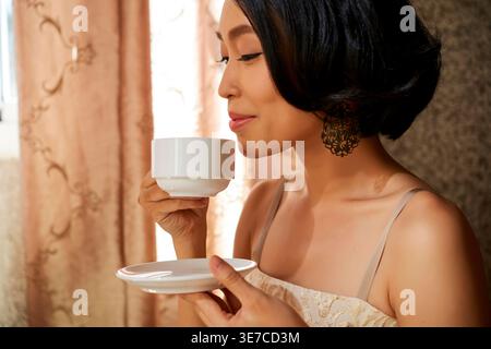 Vietnamese woman smelling aroma from coffee cup standing at home Stock Photo