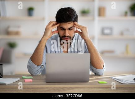 Arab man with beard touching head while sitting at desk with laptop at home Stock Photo
