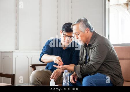 Asian middle-aged man giving medicine to his senior father sitting beside him on couch at home Stock Photo
