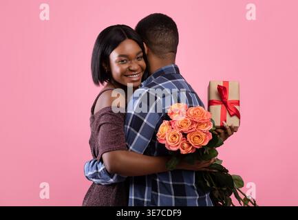 Black woman with gift box and bouquet of roses hugging her boyfriend while standing against pink background in studio Stock Photo