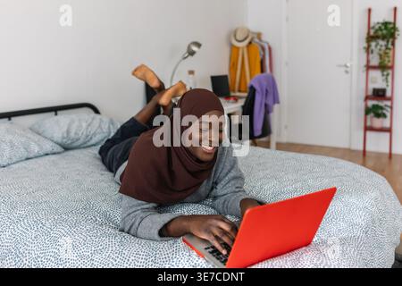 Black Muslim female teenager in brown hijab using laptop lying down on bed at home Stock Photo