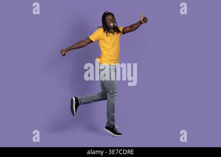 Cheerful Black man with dreads wearing yellow shirt gesturing thumbs up while jumping against purple background in studio Stock Photo