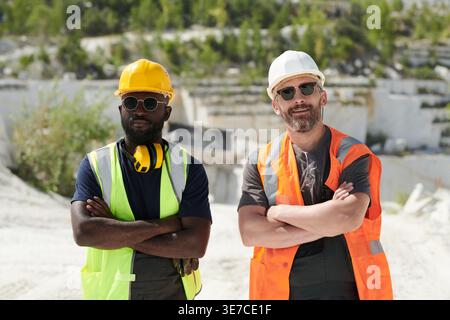 Black and caucasian male worker wearing safety vest and hard hat looking away while standing with crossed arms in construction site during sunny day Stock Photo