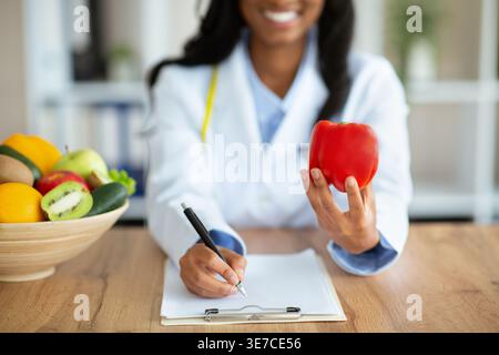 Black female nutritionist showing bell pepper writing notes sitting at desk in office Stock Photo