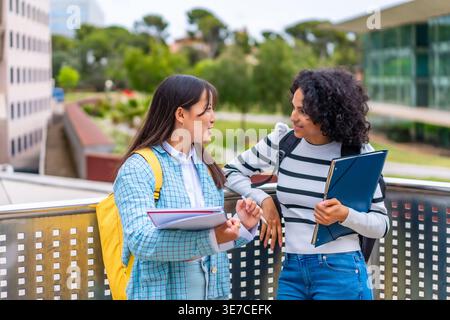 Hispanic and Asian female students with backpacks leaning on metal railings and talking to each other while standing outdoors in campus university ... Stock Photo
