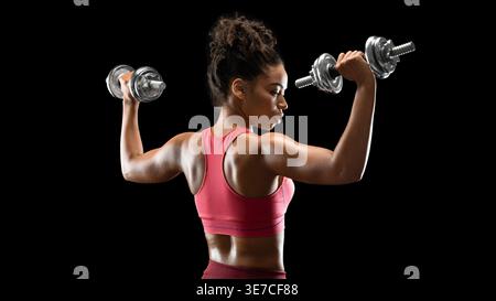 Rear view of Black woman holding dumbbells doing shoulder press while standing against black background in studio Stock Photo