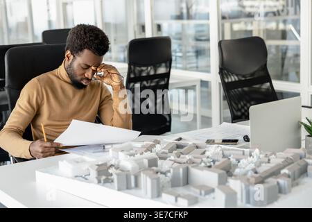 Tired African American male architect reviewing blueprints in office Stock Photo