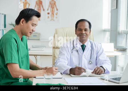 Asian male doctors in scrub suit and white lab coat with stethoscope around neck writing and sitting at table in hospital office Stock Photo