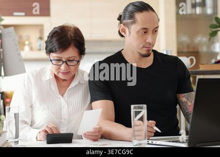 Senior mixed-race mother wearing eyeglasses computing bills on calculator with her son using laptop while sitting together at table in living room ... Stock Photo