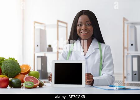 Black female nutritionist with tape measure around her neck holding tablet with blank screen sitting at table with fruits and vegetables in clinic Stock Photo