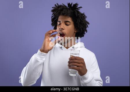 Black man with afro wearing white hoodie taking pill medicine holding glass of water while standing against purple wall in studio Stock Photo