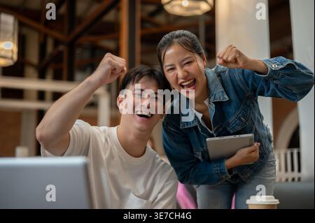 Thai male and female gesturing success with fists closed and arms raised standing and sitting in coffee shop Stock Photo