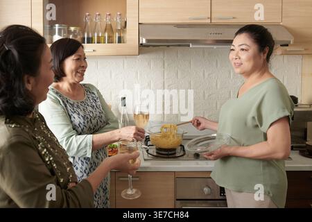 Asian senior friends holding wine glasses talking with friend cooking in kitchen at home Stock Photo