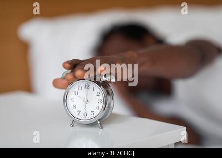 Hands of Black man turning off alarm clock on bedside table while lying on bed at home Stock Photo