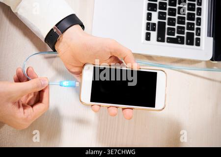 Hand of male connecting smLifestylephone to usb charging cable at desk with laptop in office Stock Photo