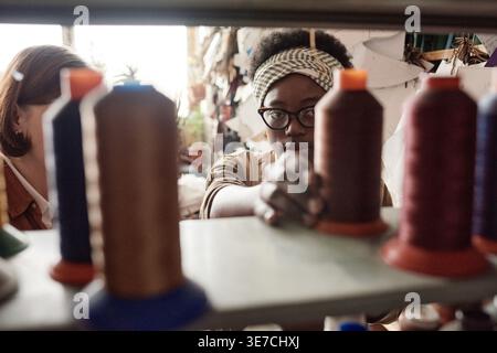 Black female tailor wearing eyeglasses standing getting threads on shelf in workshop Stock Photo