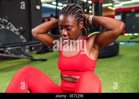 Black woman with braids in sportswear doing abdominal exercises sitting on ground in gym Stock Photo