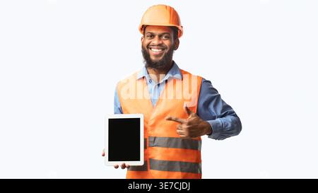 Black male worker wearing safety helmet pointing finger and showing tablet screen while standing against white background in studio Stock Photo