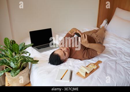Young man relaxing on bed, eating breakfast and texting friends Stock Photo