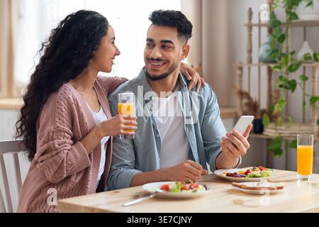 Smiling Middle Eastern couple having breakfast holding glass of juice and smartphone while sitting at table at home Stock Photo