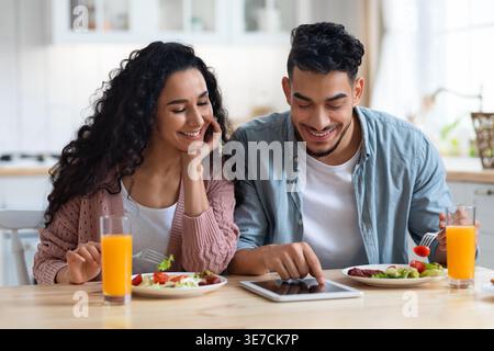 Smiling Middle Eastern couple having breakfast using tablet while sitting at table at home Stock Photo