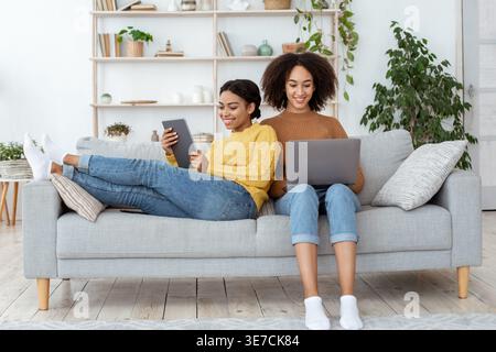 Black woman using tablet leaning on female friend using laptop while sitting on sofa in living room at home Stock Photo
