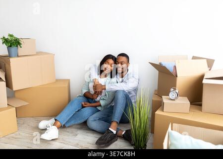 Smiling Black couple with closed eyes embracing while sitting on floor among cardboard boxes in living room at home Stock Photo