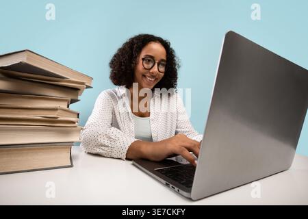 Tired Black female teenager wearing eyeglasses using laptop while sitting at table with stacks of books against blue background in studio Stock Photo