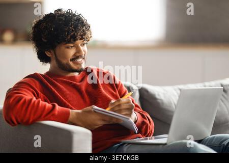 Online Education. Handsome young indian man using laptop and taking notes while sitting on couch at home, smiling eastern guy watching tutorial, lectu Stock Photo