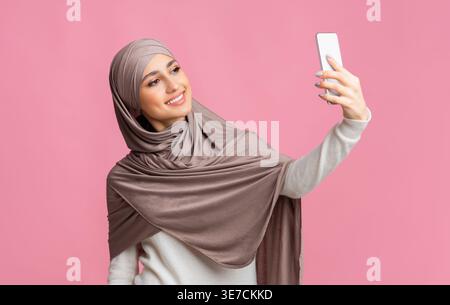 Arab woman wearing hijab taking selfie using smartphone while standing against pink background Stock Photo