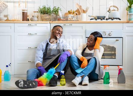 Black husband and wife wearing apron and gloves talking while sitting on floor with cleaning tools in kitchen at home Stock Photo