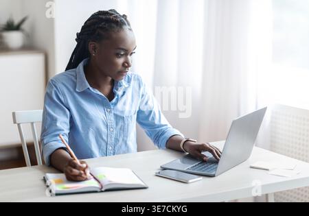 Black business woman with braids taking notes using laptop while sitting at table in home office Stock Photo