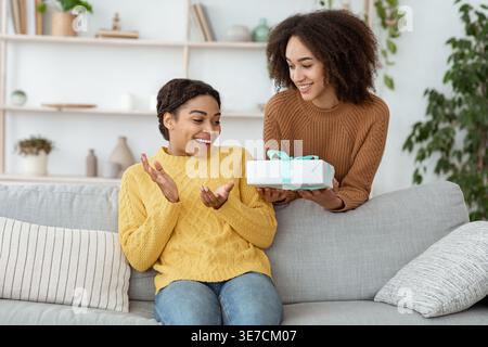 Black woman giving gift box to female friend while sitting on sofa in living room at home Stock Photo