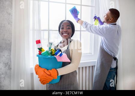Black wife wearing apron and gloves holding cleaning tools while standing near husband cleaning window in living room at home Stock Photo