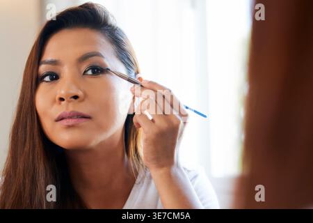 Asian woman applying eyeshadow using brush in front of mirror in living room at home Stock Photo