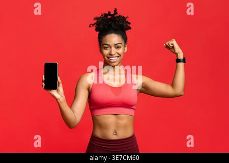 Black woman holding smLifestylephone and flexing bicep while standing against red background in studio Stock Photo