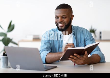 Smiling bearded Black male freelancer wearing blue long-sleeved shirt taking notes while working on laptop sitting at table in home office Stock Photo