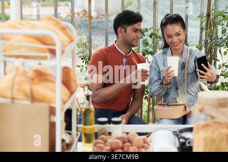 Indian man and Vietnamese woman drinking and taking selfie on smartphone while sitting on bench in an outdoor cafe Stock Photo