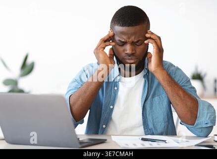 Tired bearded Black male freelancer wearing blue long-sleeved shirt massaging temples suffering from headache sitting at table with laptop and docu... Stock Photo