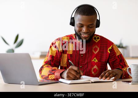 Bearded Black male freelancer wearing long-sleeved dashiki shirt listening to music on headphones writing on notebook while having videocall on lap... Stock Photo