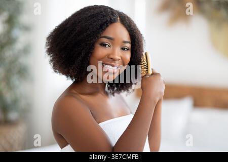 Smiling Black woman with curly hair wrapped in bath towel combing her hair with wooden brush after shower looking at mirror while standing in bedro... Stock Photo